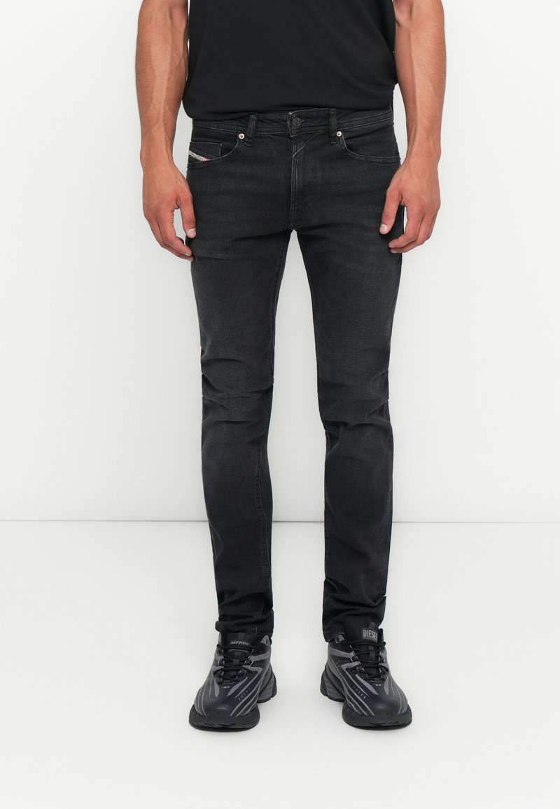 Mann, der eng anliegende schwarze Jeans und schwarze Sportschuhe trägt, steht vor einem weißen Hintergrund, die Hände entspannt an den Seiten.