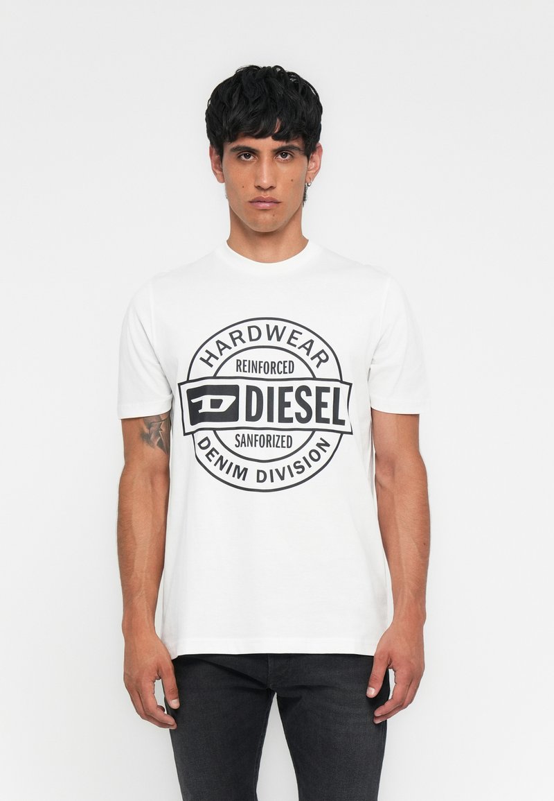 Mann trägt ein weißes T-Shirt der Marke Diesel mit schwarzem kreisförmigem Logo und Text, kombiniert mit schwarzen Jeans, stehend vor weißem Hintergrund.