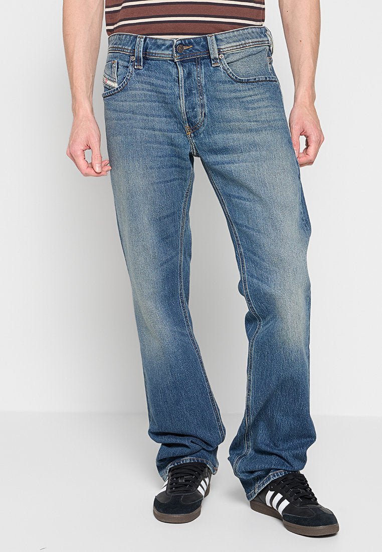Männliches Model trägt blaue, ausgebleichte gerade Jeans, schwarze Adidas-Sneaker mit weißen Streifen und ein braun gestreiftes Hemd, steht vor weißem Hintergrund.