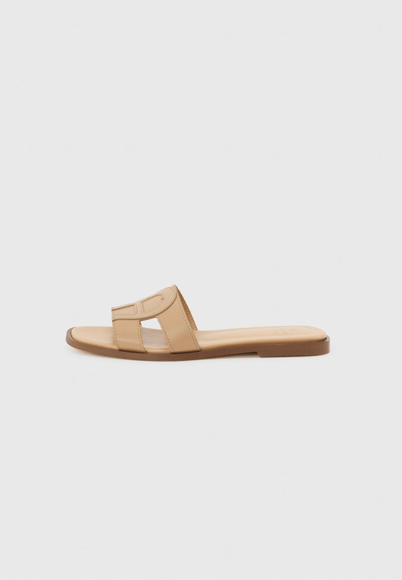 Beige flache Sandale mit breitem ausgeschnittenem Riemen und quadratischer Spitze, seitlich vor hellem grauem Hintergrund gezeigt.