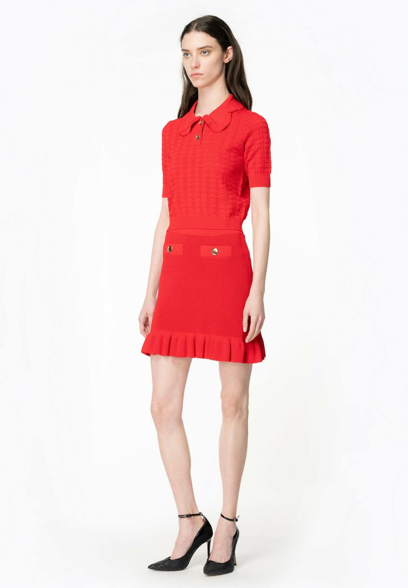 Rotes gestricktes Kleid mit einem kurzärmeligen Polokragen, dekorativen Knöpfen und einem Rüschensaum; kombiniert mit schwarzen High Heels.
