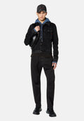 Schwarze Denim-Jacke mit Fronttaschen, über einem blauen Hoodie getragen. Schwarze Hose und Sneakers vervollständigen den Look, mit einer lädierten schwarzen Mütze.