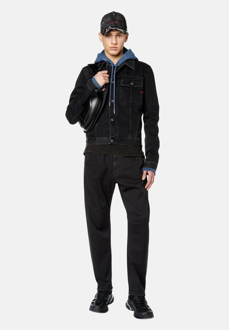 Schwarze Denim-Jacke mit Fronttaschen, über einem blauen Hoodie getragen. Schwarze Hose und Sneakers vervollständigen den Look, mit einer lädierten schwarzen Mütze.