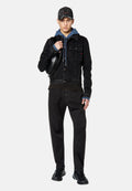 Schwarze Denim-Jacke mit Fronttaschen, über einem blauen Hoodie getragen. Schwarze Hose und Sneakers vervollständigen den Look, mit einer lädierten schwarzen Mütze.