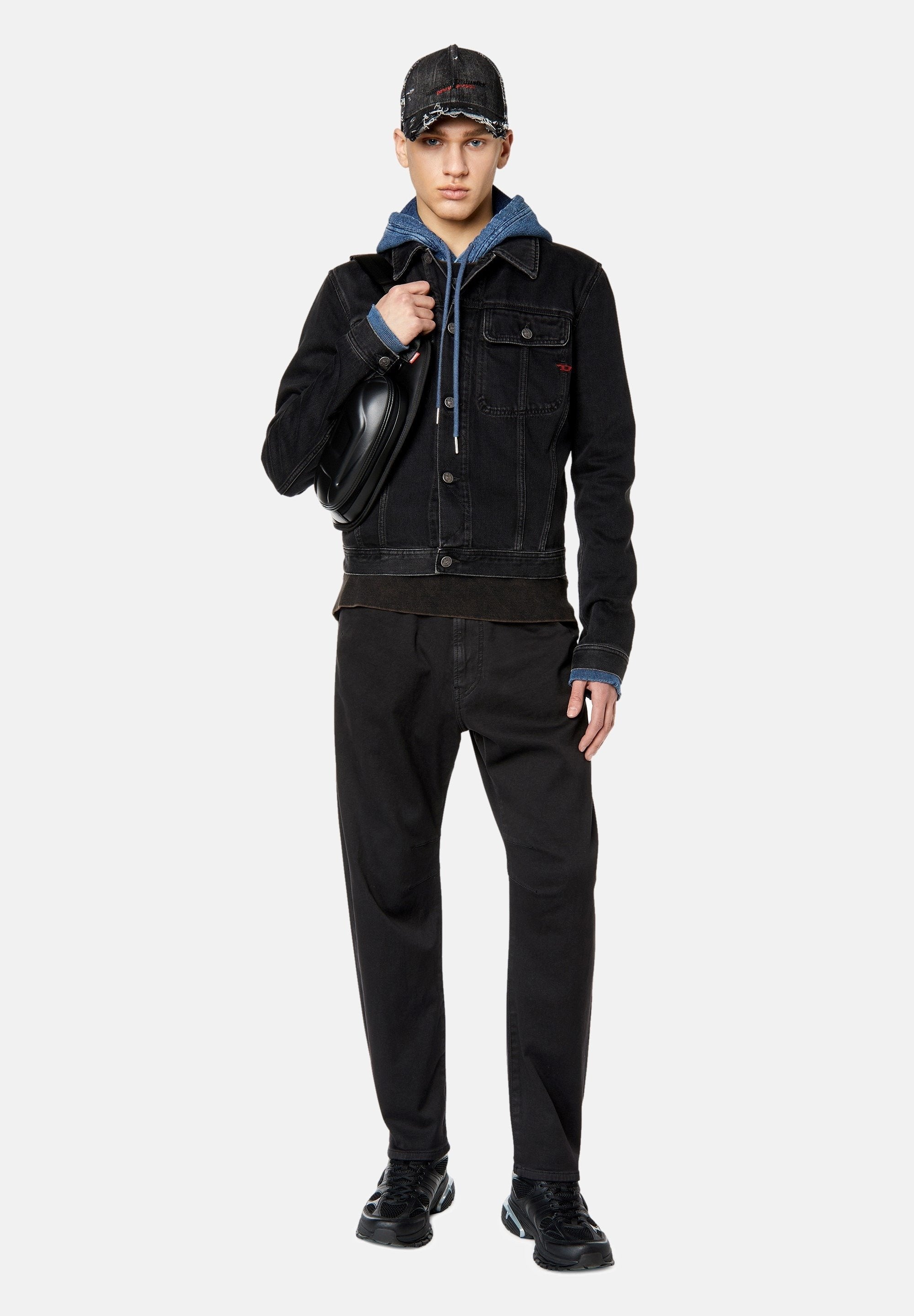 Schwarze Denim-Jacke mit Fronttaschen, über einem blauen Hoodie getragen. Schwarze Hose und Sneakers vervollständigen den Look, mit einer lädierten schwarzen Mütze.