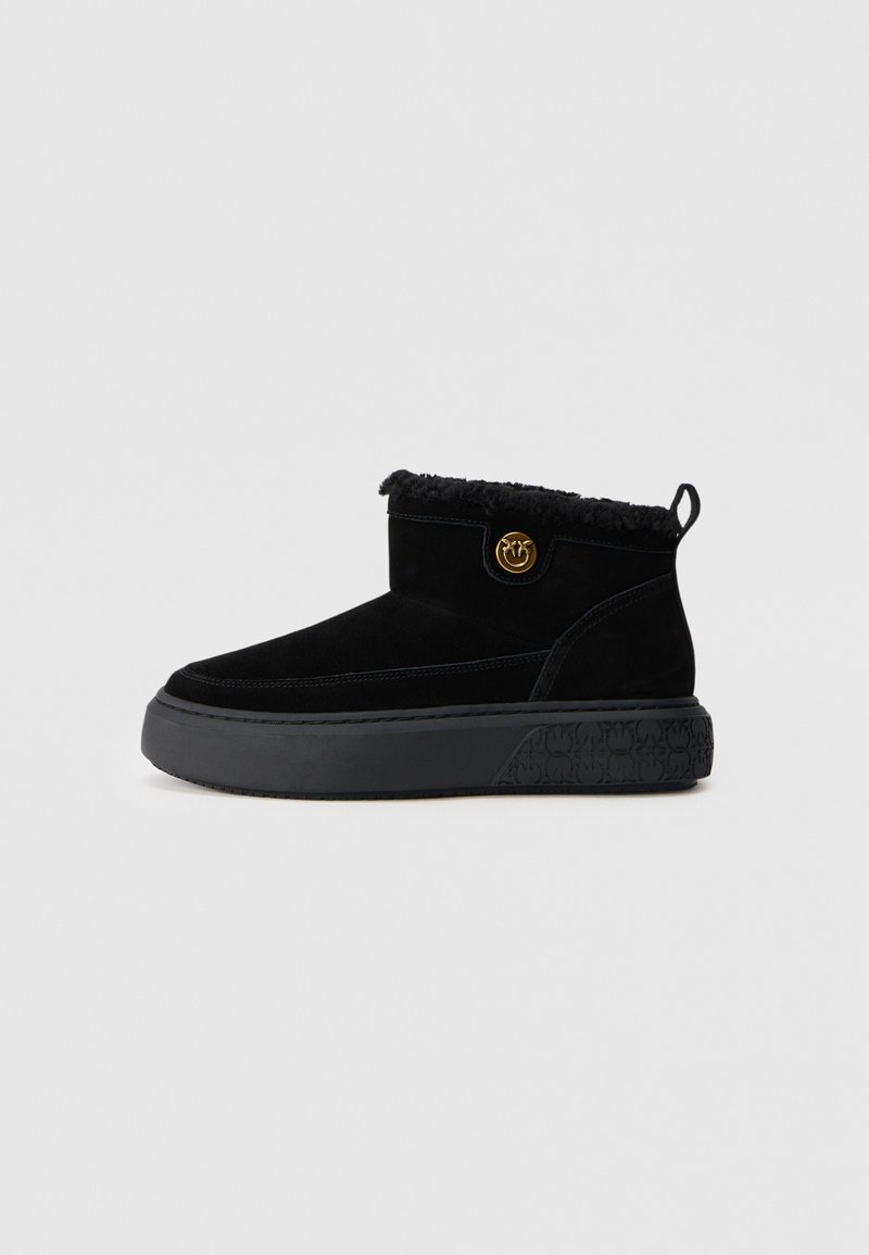 Schwarze Knöchelstiefel mit einem Obermaterial aus Wildleder, einer weichen Fellverzierung, einem runden goldenen Emblem und einer dicken strukturierten Gummisohle.