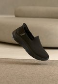 Schwarze Slipper mit Mesh-Obermaterial, strukturiertem Absatz und flexibler Sohle. Leichtes Design. Hintergrund mit sanften, neutralen Farbtönen.