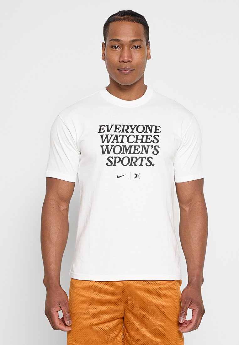 Mann trägt ein weißes T-Shirt mit der Aufschrift "JEDER SCHAUF FRAUENSPORT" und orangefarbene Sportshorts, steht vor einem schlichten Hintergrund.