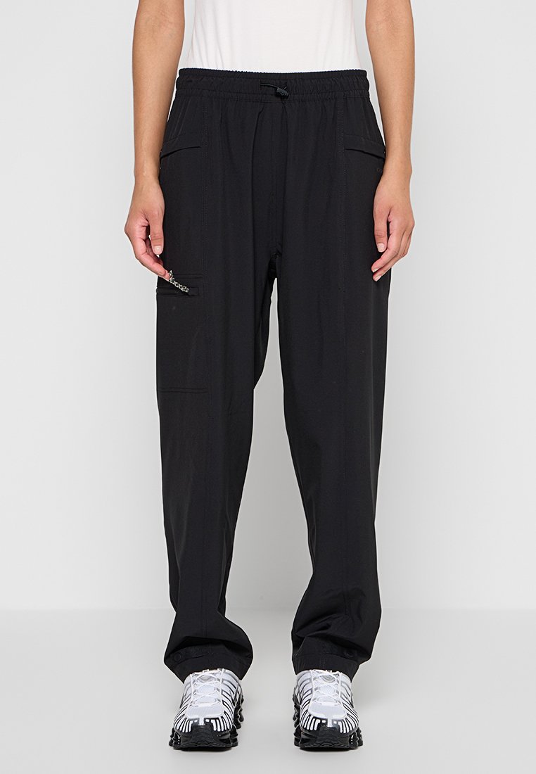 Person, der schwarze Cargo-Hosen mit elastischem Bund und Taschen trägt, kombiniert mit weißen und schwarzen Sportschuhen, steht vor einem einfarbigen Hintergrund.