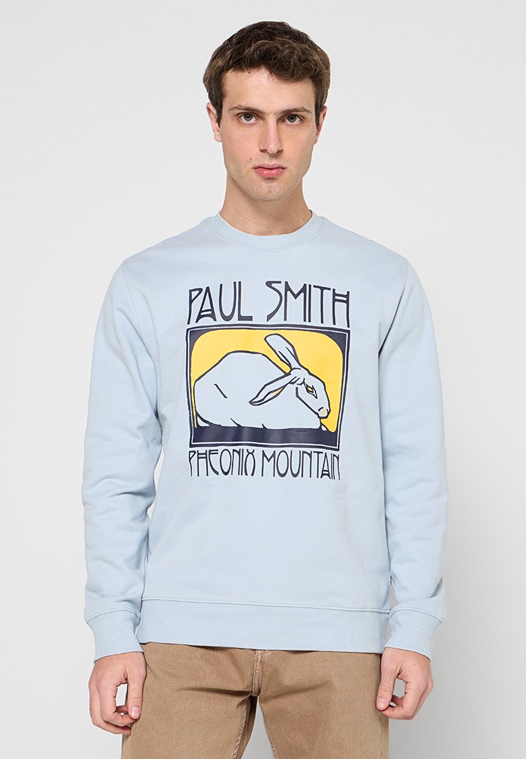 Junger Mann trägt ein hellblaues Sweatshirt mit den Aufschriften „Paul Smith“ und „Phoenix Mountain“ über und unter einer Illustration eines weißen Kaninchens.