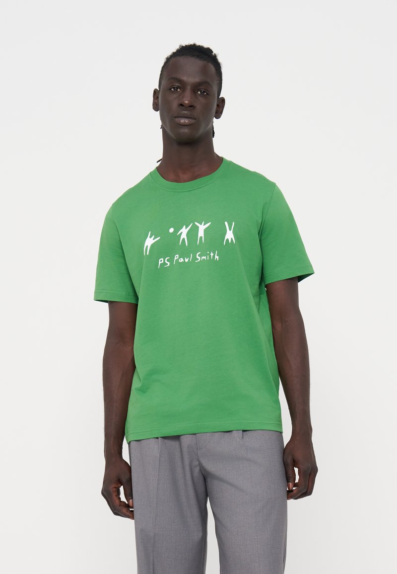 Mann trägt ein leuchtend grünes T-Shirt mit weißen Strichmännchen-Illustrationen und dem Schriftzug „PS Paul Smith“, kombiniert mit grauer Hose.