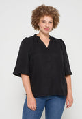 Schwarze Bluse mit strukturiertem Finish, gekennzeichnet durch kurze, ausgestellte Ärmel und einen V-Ausschnitt. Kombiniert mit blauen Jeans, vor einem neutralen Hintergrund präsentiert.