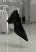 Schwarzer High-Heel-Schuh mit spitzem Zehenbereich und blockiertem Absatz, aus strukturiertem Leder, über einem glatten Boden schwebend.