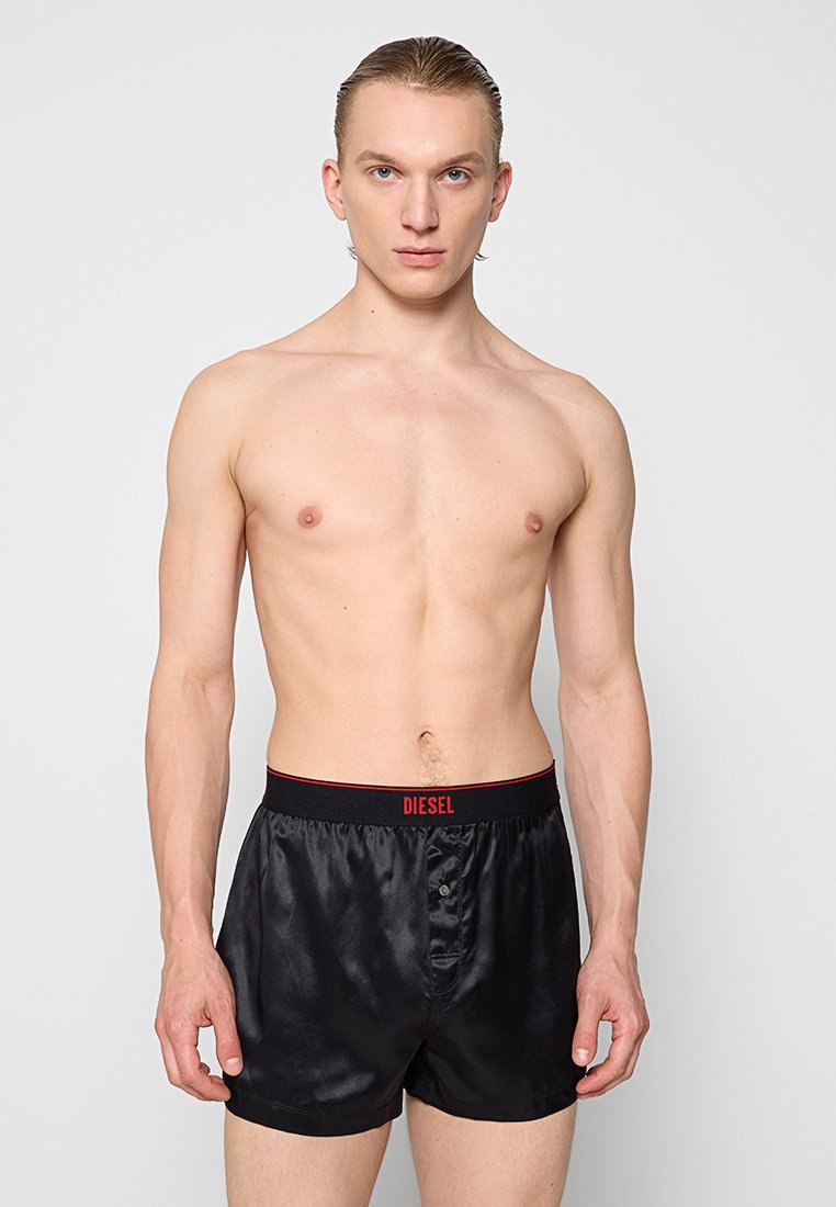 Schwarze Satin-Boxershorts mit rotem Bund, der das "DIESEL"-Logo trägt. Sie sind mit einem Knopfverschluss und einer glatten Textur gestaltet.