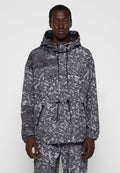 Mann in grauer und schwarzer Camouflage-Jacke mit Kapuze und passenden Hosen, der vor einem schlichten, hellen Hintergrund steht und nach vorne schaut.