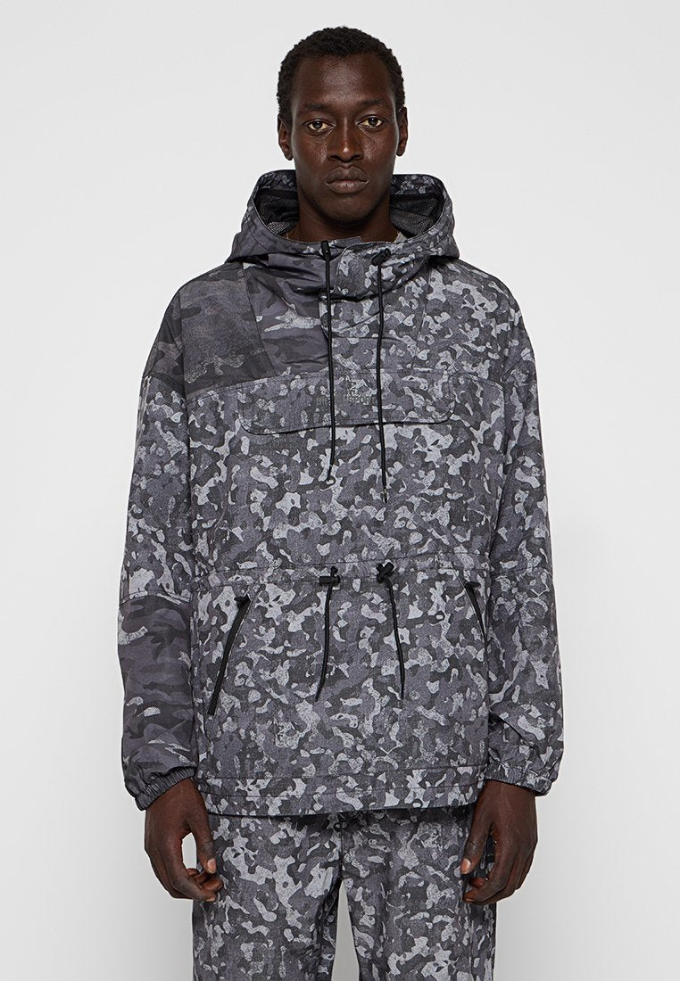 Mann in grauer und schwarzer Camouflage-Jacke mit Kapuze und passenden Hosen, der vor einem schlichten, hellen Hintergrund steht und nach vorne schaut.
