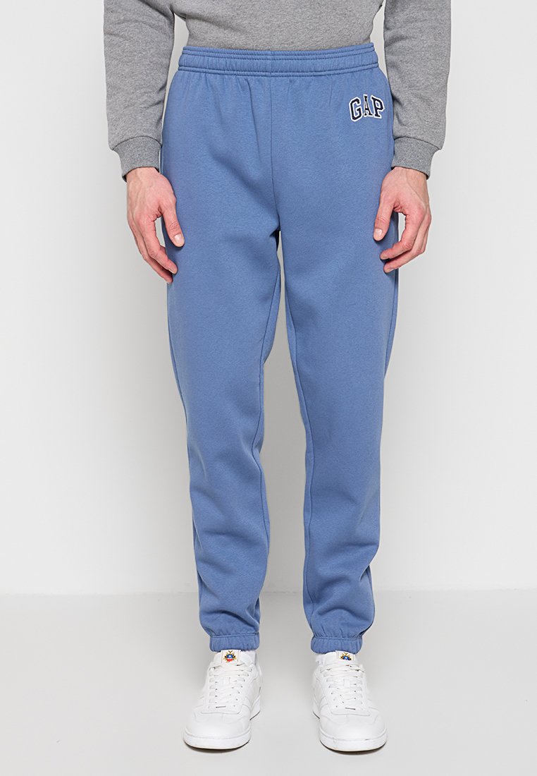 Mann trägt blaue GAP-Sweathosen, weiße Turnschuhe und einen grauen Pullover, steht vor einem schlichten weißen Hintergrund.
