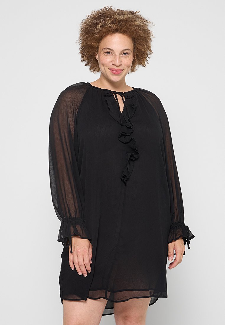 Schwarzes Kleid mit langen, transparenten Ärmeln, rüschigem Vorderteil und rundem Ausschnitt. Verfügt über einen Bindebandverschluss am Ausschnitt und elastische Bündchen.