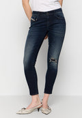 Eine Frau trägt dunkle blaue enge Jeans mit einem aufgerissenen Knie und beigen, flachen Slingback-Schuhen. Sie steht vor einem weißen Hintergrund.