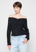 Frau mit kurzem braunem Haar, die einen schwarzen schulterfreien Rippenpullover und hellblaue hoch taillierte Jeans vor einem einfarbigen Hintergrund trägt.