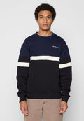 Junger Mann mit lockigem Haar, der einen dunkelblauen, schwarzen und weißen gestreiften Sweatshirt und beige Hosen trägt, stehend vor einem schlichten Hintergrund.