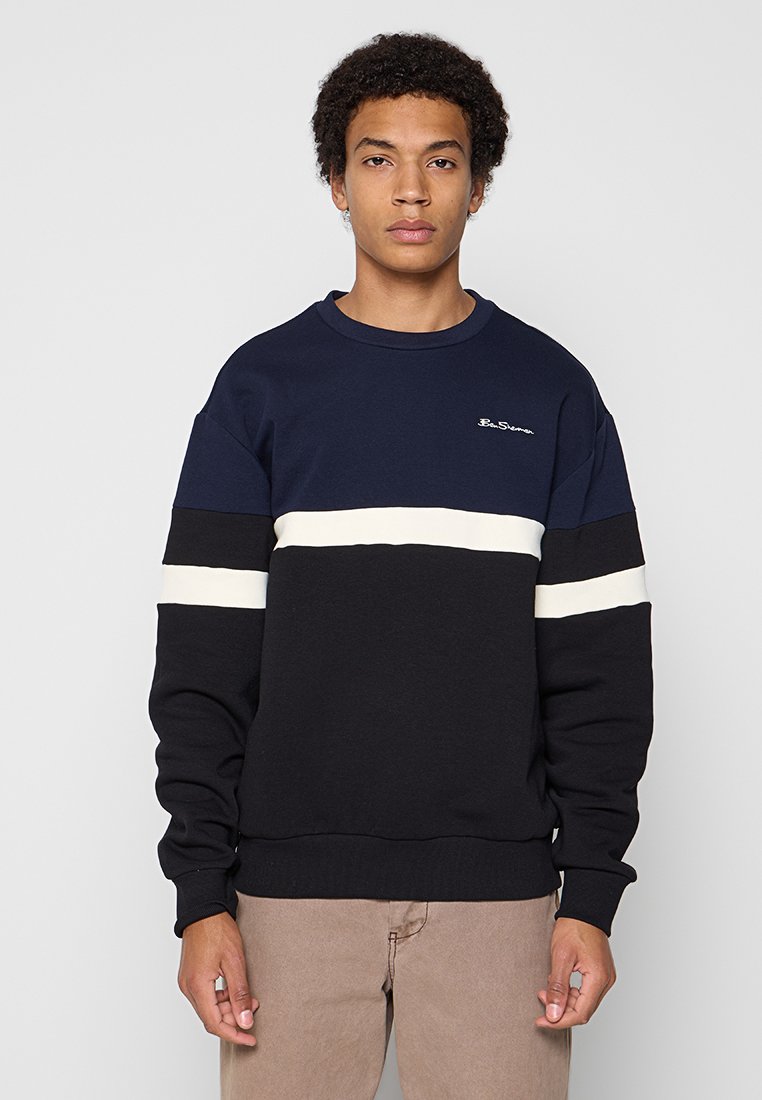 Junger Mann mit lockigem Haar, der einen dunkelblauen, schwarzen und weißen gestreiften Sweatshirt und beige Hosen trägt, stehend vor einem schlichten Hintergrund.
