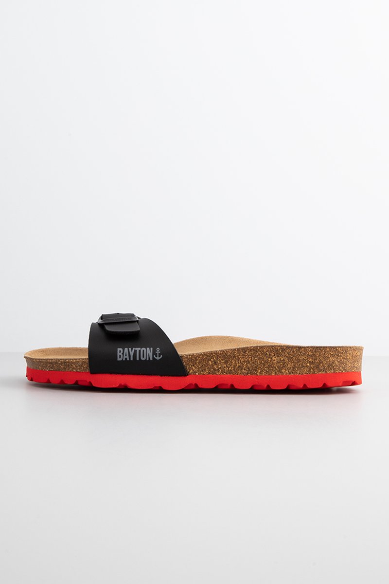 Schwarze Rutschsandale mit einem Korkfußbett und roter Gummisohle. Verfügt über einen schwarzen Riemen mit dem "BAYTON"-Logo und einer sicheren Schnalle.