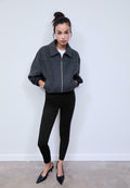 Graue Zip-Jacke mit strukturiertem Kragen, kombiniert mit schwarzen, eng anliegenden Hosen und spitzen schwarzen Absätzen, steht vor einem neutralen Hintergrund.