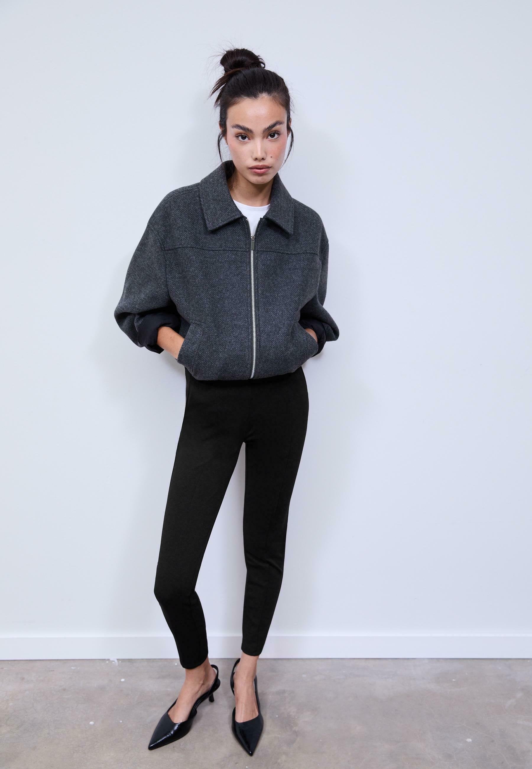 Graue Zip-Jacke mit strukturiertem Kragen, kombiniert mit schwarzen, eng anliegenden Hosen und spitzen schwarzen Absätzen, steht vor einem neutralen Hintergrund.