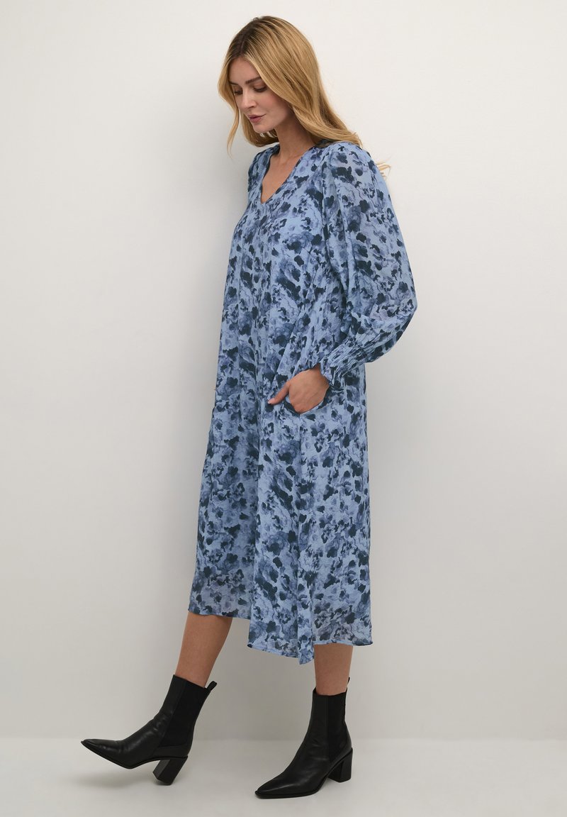 Langärmliges, V-Ausschnitt-Kleid in Hellblau mit dunkelblauen Blumenmustern, fließender Form, seitlichen Taschen, kombiniert mit schwarzen Ankle-Boots.