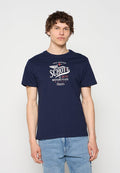 Junger Mann trägt ein dunkelblaues T-Shirt mit „Schott Motorcycles“-Grafik und hellblaue Jeans, steht vor einem einfarbigen weißen Hintergrund.