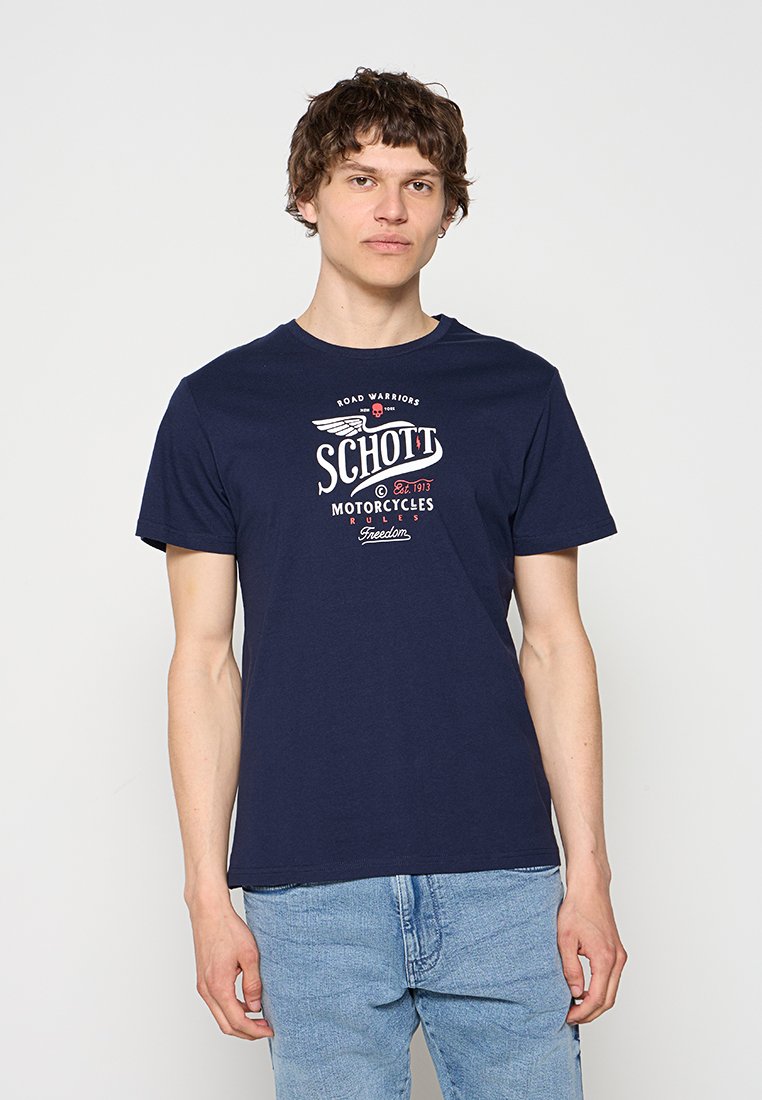 Junger Mann trägt ein dunkelblaues T-Shirt mit „Schott Motorcycles“-Grafik und hellblaue Jeans, steht vor einem einfarbigen weißen Hintergrund.