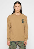 Langärmliges, beige Oberteil mit rundem Ausschnitt und grünem Grafik-Logo auf der linken Brust. Kombiniert mit Camouflage-Cargohosen.