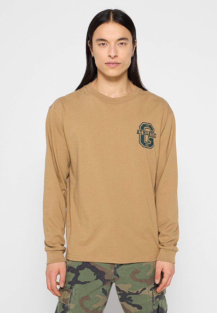 Langärmliges, beige Oberteil mit rundem Ausschnitt und grünem Grafik-Logo auf der linken Brust. Kombiniert mit Camouflage-Cargohosen.