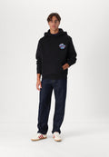 Junger Mann mit schwarzem Kapuzenpullover mit Brustgrafik, dunkler Jeans und weißen Turnschuhen mit roten Streifen, der vor einem weißen Hintergrund steht.