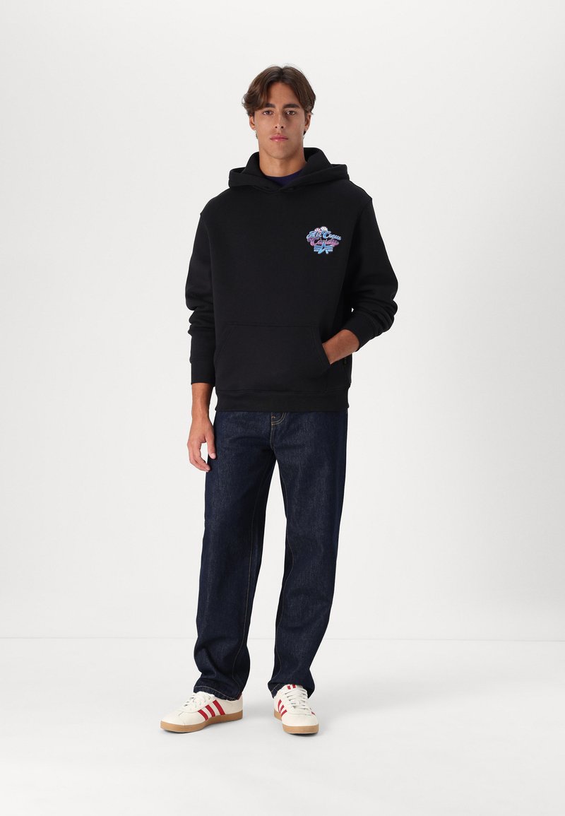 Junger Mann mit schwarzem Kapuzenpullover mit Brustgrafik, dunkler Jeans und weißen Turnschuhen mit roten Streifen, der vor einem weißen Hintergrund steht.