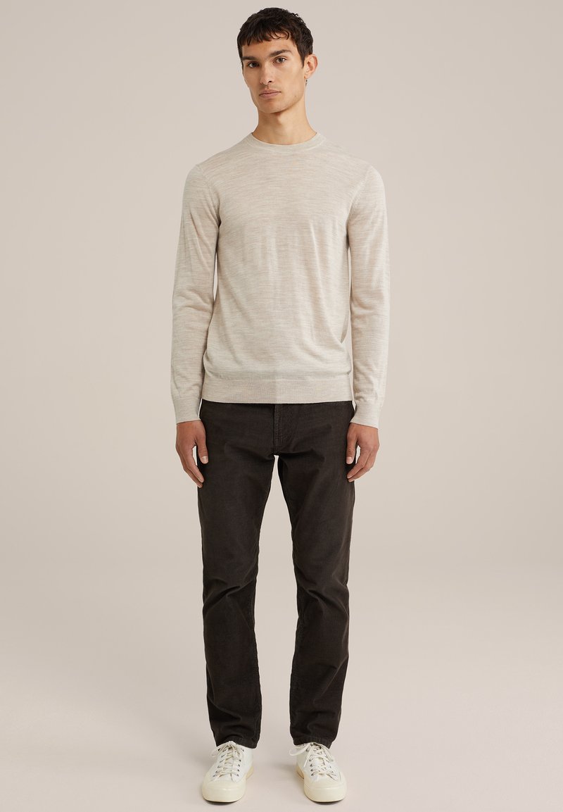 Helle beige Langarmpullover mit Rundhalsausschnitt, strukturierter Stoff. Getragen mit dunklen Hosen und weißen Sneakers. Modell steht, neutraler Hintergrund.