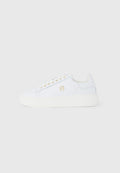 Weißer Sneaker mit niedrigem Schaft, dicker Sohle, goldenen Ösen und einem kleinen goldenen Emblem an der Seite, vor schlichtem, hellem Hintergrund.