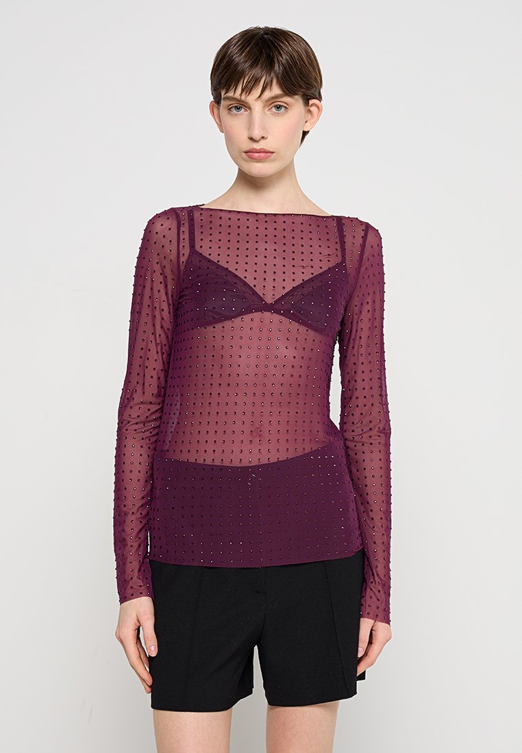 Durchsichtige lila Langarm-Bluse mit Punktmuster, über einem passenden Bralette getragen. Kombiniert mit schwarzen hochsitzienden Shorts.