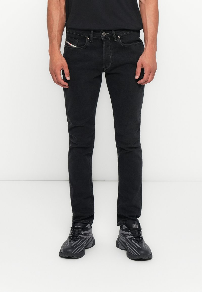 Mann, der vor einem weißen Hintergrund steht, trägt schmale schwarze Jeans und schwarz-graue Sportschuhe, die Hände entspannt an den Seiten.
