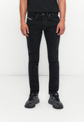 Mann, der vor einem weißen Hintergrund steht, trägt schmale schwarze Jeans und schwarz-graue Sportschuhe, die Hände entspannt an den Seiten.