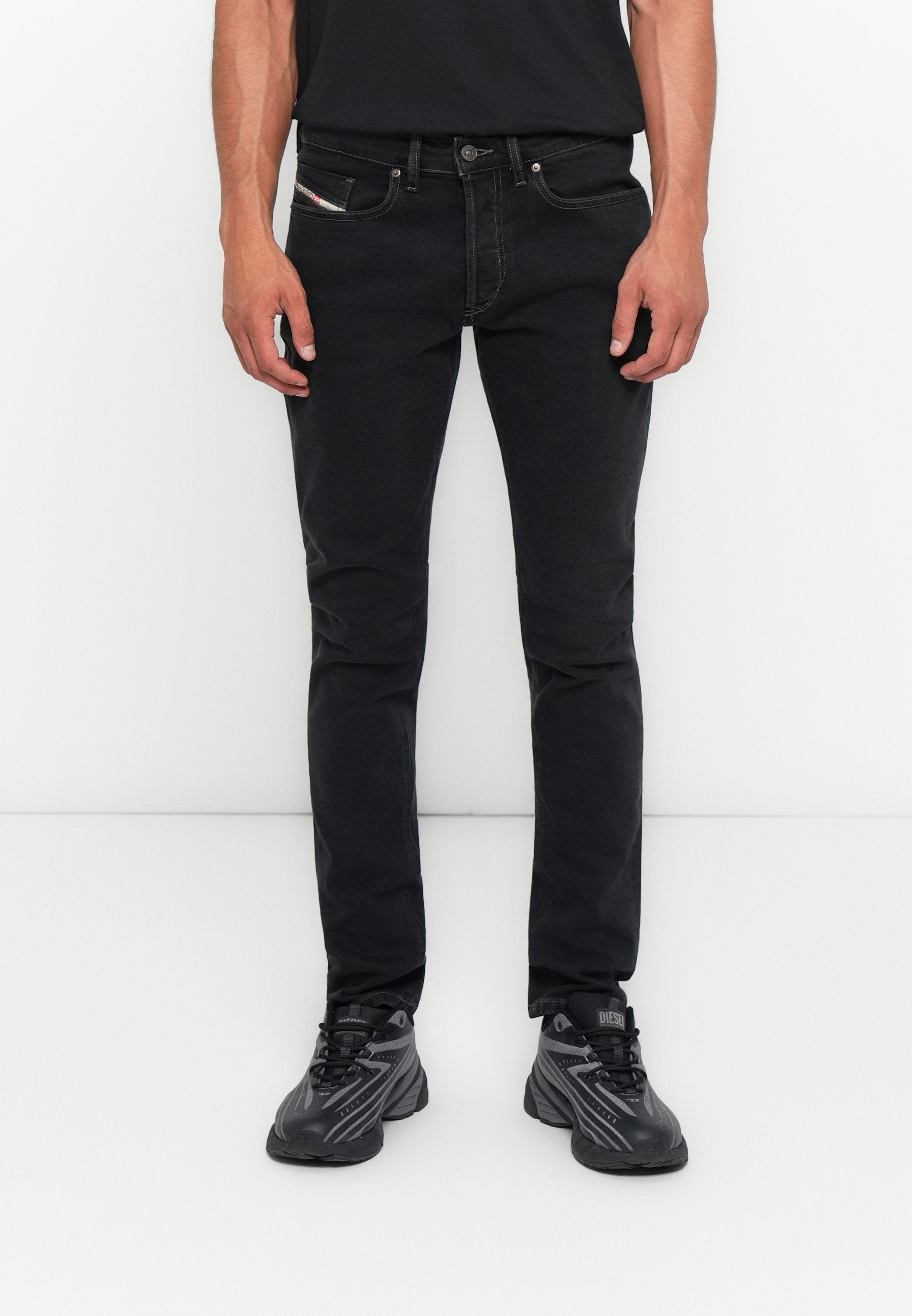 Mann, der vor einem weißen Hintergrund steht, trägt schmale schwarze Jeans und schwarz-graue Sportschuhe, die Hände entspannt an den Seiten.