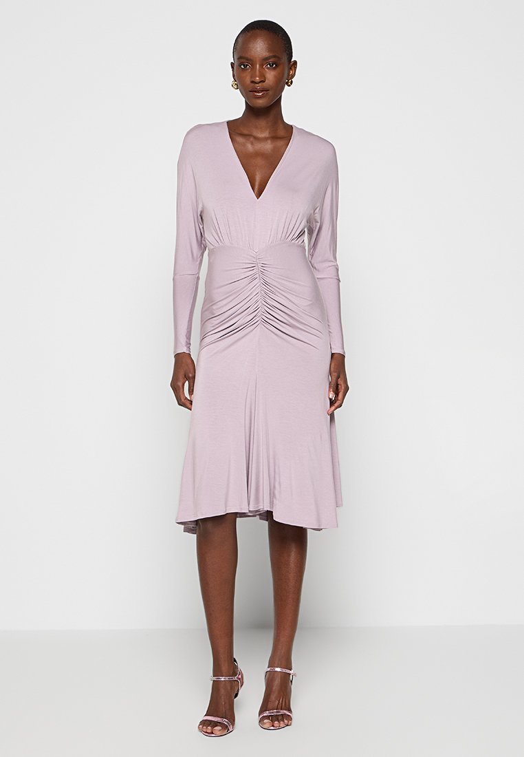 Fliederfarbenes langärmliges Kleid mit tiefem V-Ausschnitt, geraffter Vorderseite und ausgestelltem Saum. Weicher Stoff mit glatter Textur. Kombiniert mit Riemchen-Heels.
