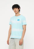 Junger Mann mit lockigem dunklem Haar, der ein hellblau-weiß gestreiftes T-Shirt und helle Jeans trägt, steht vor einem schlichten weißen Hintergrund.