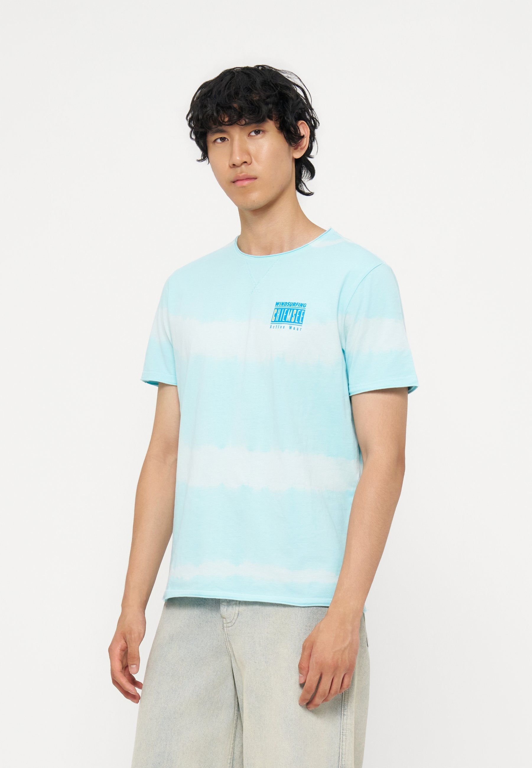 Junger Mann mit lockigem dunklem Haar, der ein hellblau-weiß gestreiftes T-Shirt und helle Jeans trägt, steht vor einem schlichten weißen Hintergrund.