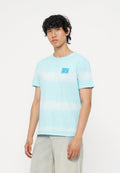 Junger Mann mit lockigem dunklem Haar, der ein hellblau-weiß gestreiftes T-Shirt und helle Jeans trägt, steht vor einem schlichten weißen Hintergrund.