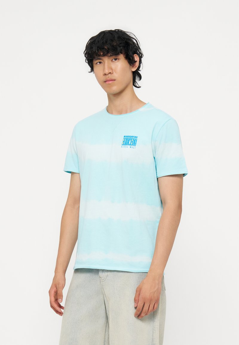 Junger Mann mit lockigem dunklem Haar, der ein hellblau-weiß gestreiftes T-Shirt und helle Jeans trägt, steht vor einem schlichten weißen Hintergrund.