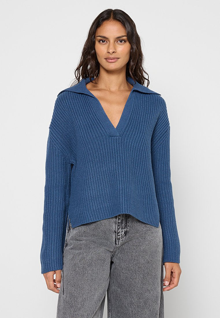 Blauer Strickpullover mit V-Ausschnitt und weiter Kragen. Mit strukturierter Rippenoptik und kombiniert mit grauen hoch geschnittenen Jeans.