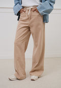 Person trägt beige Hosen mit weitem Bein und weißem Kordelzug, weiße Sneaker, ein weißes Oberteil und eine hellblaue Jeansjacke, die Hände in den Taschen.