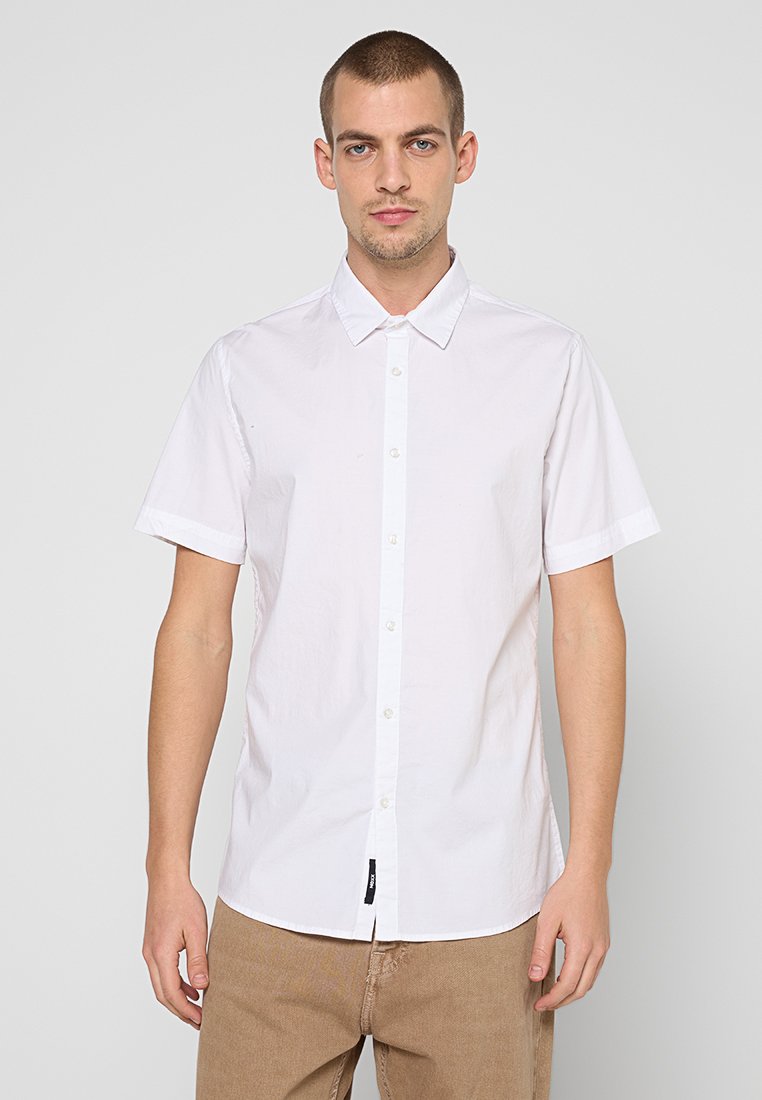 Weißes kurzärmeliges Hemd mit Button-Down-Kragen, versehen mit einer Frontblende und einer strukturierten Oberfläche. Kombiniert mit hellbraunen Hosen.
